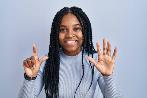 Αφροαμερικανή γυναίκα στέκεται πάνω από το μπλε φόντο δείχνει και δείχνει προς τα πάνω με τα δάχτυλα νούμερο επτά, ενώ χαμογελά αυτοπεποίθηση και χαρούμενος.  - Φωτογραφία, εικόνα