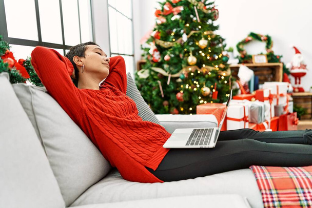 Jeune femme hispanique détendue avec les mains sur la tête à l'aide d'un ordinateur portable assis près de l'arbre de Noël à la maison - Photo, image