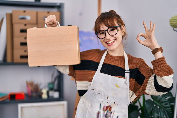 Молодая красивая художница с деревянным чехлом делает хорошо знак с пальцами, улыбаясь дружелюбный жест отличный символ  - Фото, изображение