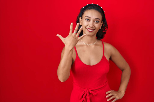 Young hispanic woman standing over red background showing and pointing up with fingers number five while smiling confident and happy.  - Foto, immagini