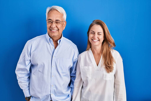Middle age hispanic couple standing over blue background winking looking at the camera with sexy expression, cheerful and happy face.  - Foto, afbeelding
