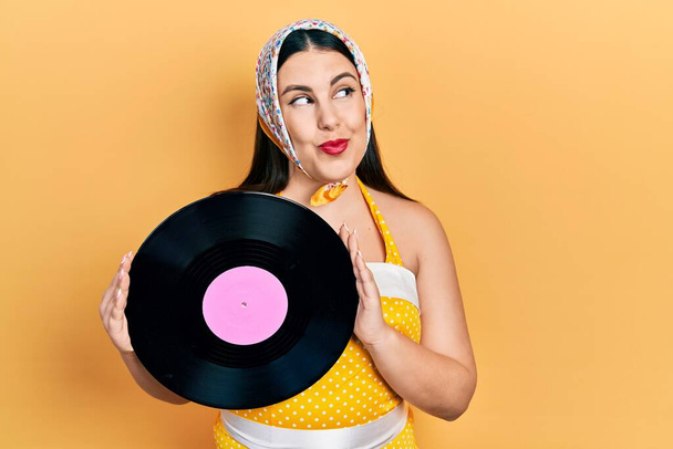 Young hispanic woman wearing pin up style holding vinyl disc smiling looking to the side and staring away thinking.  - Fotoğraf, Görsel