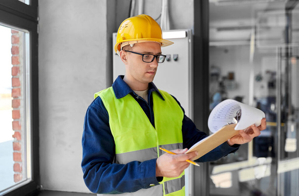 construction business and building concept - male electrician or worker in helmet and safety west with papers on clipboard and pencil at electric board - Fotó, kép