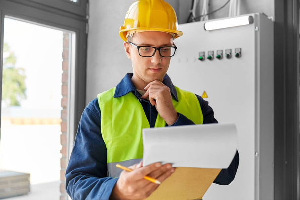 construction business and building concept - male electrician or worker in helmet and safety west with papers on clipboard and pencil at electric board - Foto, imagen
