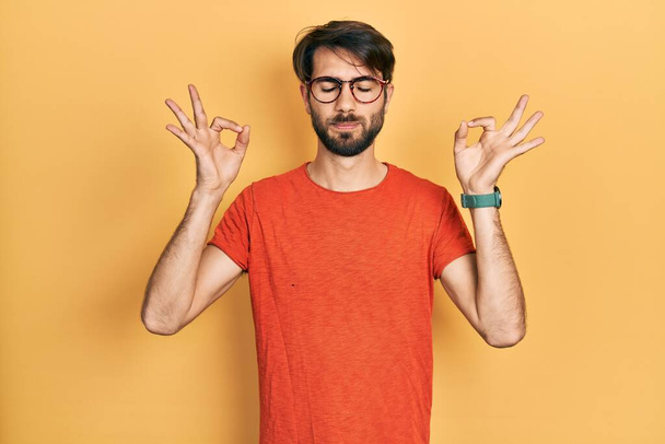 Jeune homme hispanique portant des vêtements et des lunettes décontractés se détendre et sourire les yeux fermés en faisant geste de méditation avec les doigts. concept de yoga.  - Photo, image