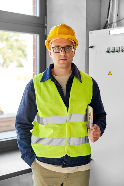 construction business and building concept - male electrician or worker in helmet and safety west with papers on clipboard and pencil at electric board - Fotoğraf, Görsel