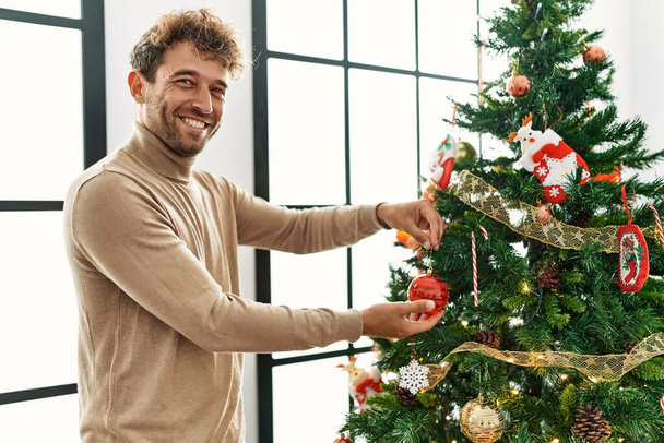 Junger hispanischer Mann lächelt selbstbewusst Weihnachtsbaum zu Hause schmücken - Foto, Bild