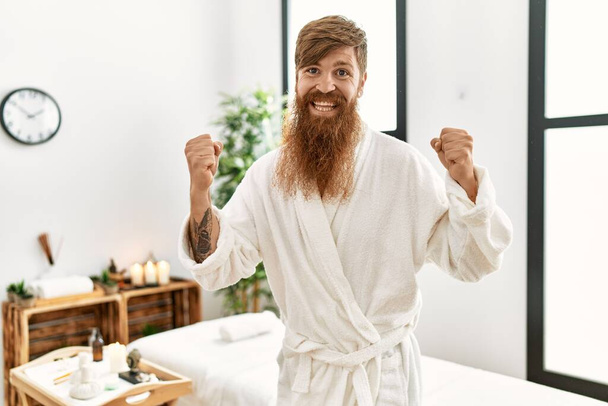 Redhead man with long beard wearing bathrobe at wellness spa screaming proud, celebrating victory and success very excited with raised arms  - Foto, afbeelding