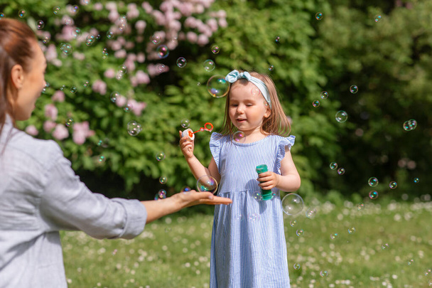 family, motherhood and people concept - happy mother with little daughter blowing soap bubbles at summer park or garden - Fotoğraf, Görsel
