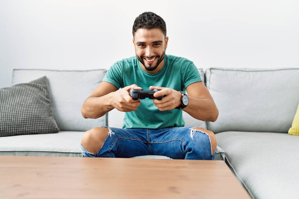 Hombre árabe joven jugando videojuego sentado en el sofá en casa - Foto, imagen