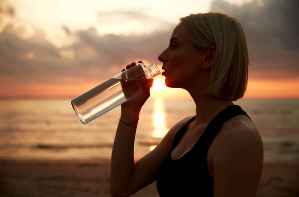fitness, sport, and healthy lifestyle concept - woman drinking water from bottle on beach over sunset - Photo, Image