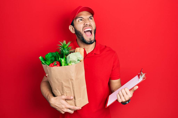 Hispanic man with beard wearing courier uniform with groceries from supermarket and clipboard angry and mad screaming frustrated and furious, shouting with anger looking up.  - Photo, image