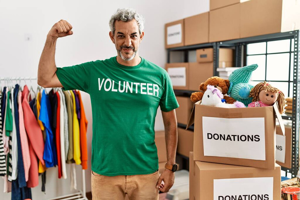 Middle age hispanic man wearing volunteer t shirt at donations stand strong person showing arm muscle, confident and proud of power  - Photo, Image