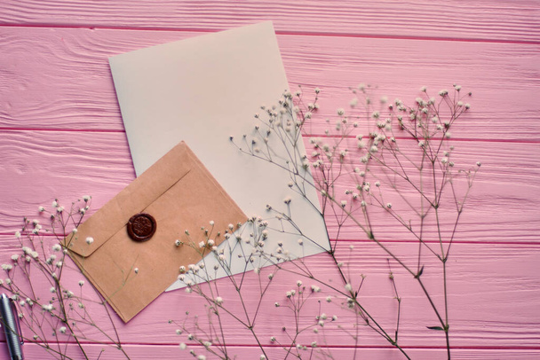 Φάκελος με λευκό χαρτί και ανοιξιάτικα λουλούδια. Ροζ ξύλινο γραφείο φόντο. - Φωτογραφία, εικόνα
