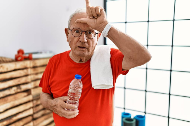 Senior man wearing sportswear and towel at the gym making fun of people with fingers on forehead doing loser gesture mocking and insulting.  - 写真・画像