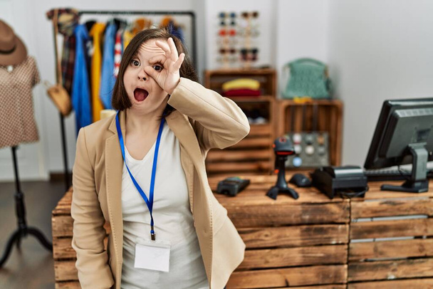 Young down syndrome woman working as manager at retail boutique doing ok gesture shocked with surprised face, eye looking through fingers. unbelieving expression.  - Foto, Imagen