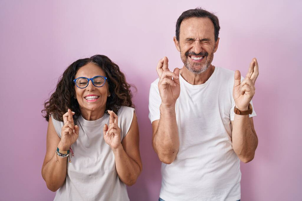 Middle age hispanic couple together over pink background gesturing finger crossed smiling with hope and eyes closed. luck and superstitious concept.  - Photo, image