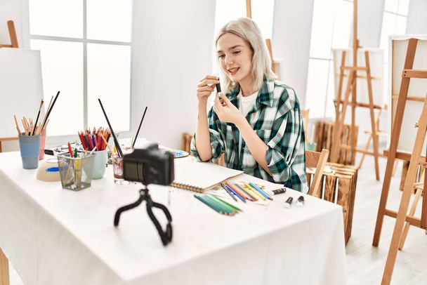 Jong kunstenares studente meisje glimlachend gelukkig schilderij. Zittend op een bureau om kunstlessen op te nemen in een kunststudio. - Foto, afbeelding