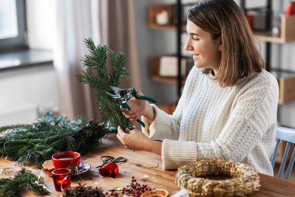 winter holidays, diy and hobby concept - happy smiling woman with fir branches and pruner making christmas wreath at home - Фото, изображение