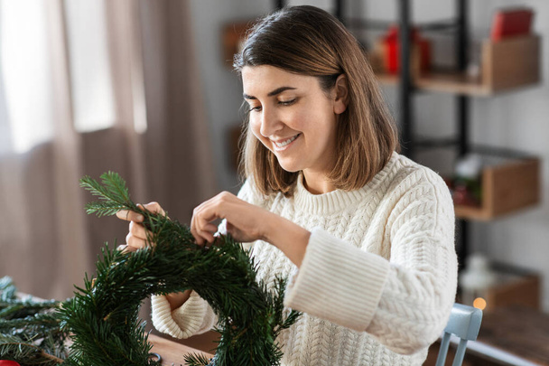 wintervakantie, diy en hobby concept - vrolijke glimlachende vrouw met sparren takken maken kerstkrans thuis - Foto, afbeelding