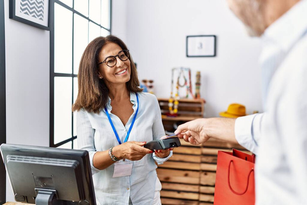 Mężczyzna i kobieta w średnim wieku uśmiechający się pewnie płacąc kartą kredytową i telefonem z danymi w sklepie odzieżowym - Zdjęcie, obraz