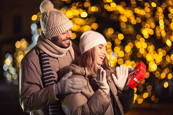 zimní prázdniny a lidé koncept - šťastný usměvavý pár s dárkem přes vánoční světla ve večerních hodinách - Fotografie, Obrázek