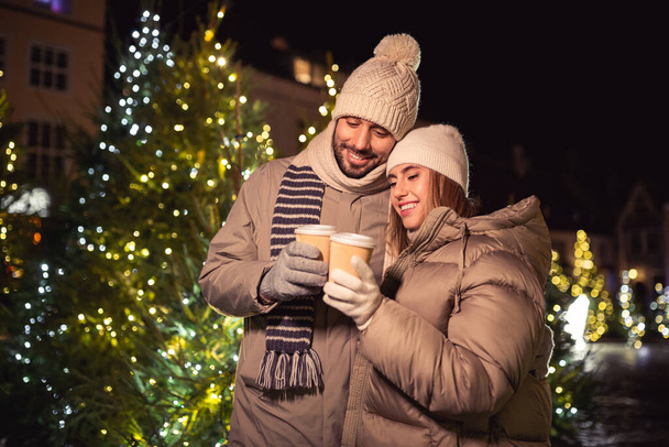 winter holidays, hot drinks and people concept - happy young couple with takeaway coffee cups over christmas tree lights in evening city - Foto, afbeelding