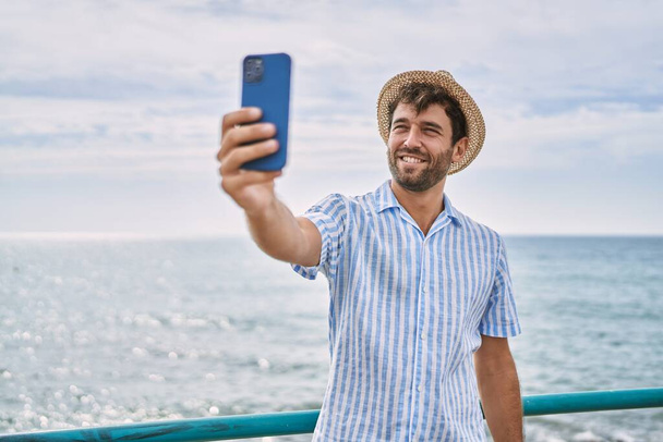 Молодой латиноамериканец улыбается счастливо делая селфи возле смартфона на пляже - Фото, изображение