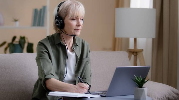 Senior businesswoman in headphones sitting on couch listen online training webinar distant e-learning 60s woman business class video chat call conference on laptop computer remote working writing note - Zdjęcie, obraz