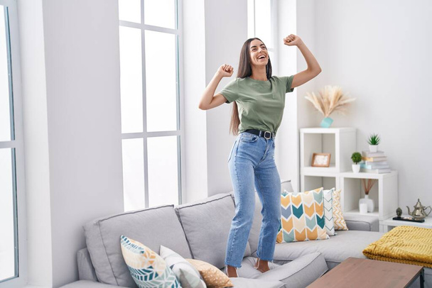 Молодая красивая латиноамериканка улыбается уверенно танцуя дома на диване - Фото, изображение