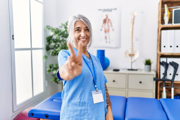 Middle age grey-haired woman wearing physiotherapist uniform at medical clinic smiling looking to the camera showing fingers doing victory sign. number two.  - Fotó, kép
