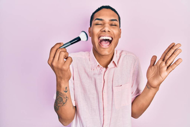 Young african american guy holding makeup brush celebrating achievement with happy smile and winner expression with raised hand  - Фото, изображение
