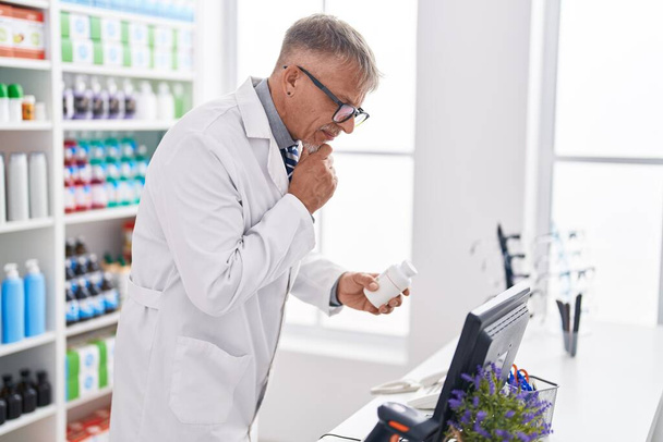 Μεσήλικας γκριζομάλλης φαρμακοποιός κρατώντας χάπια μπουκάλι χρησιμοποιώντας υπολογιστή στο εργαστήριο - Φωτογραφία, εικόνα