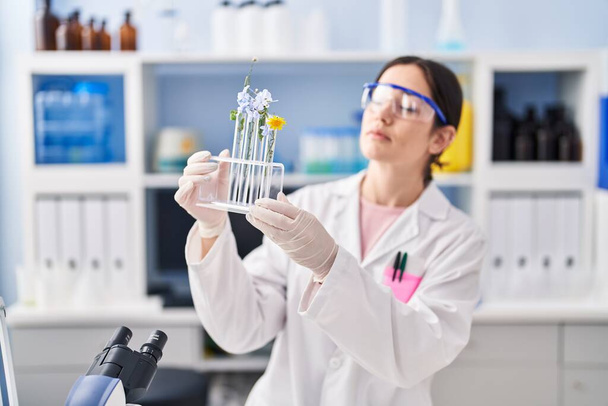 Jeune femme portant l'uniforme scientifique tenant des éprouvettes avec des fleurs au laboratoire - Photo, image