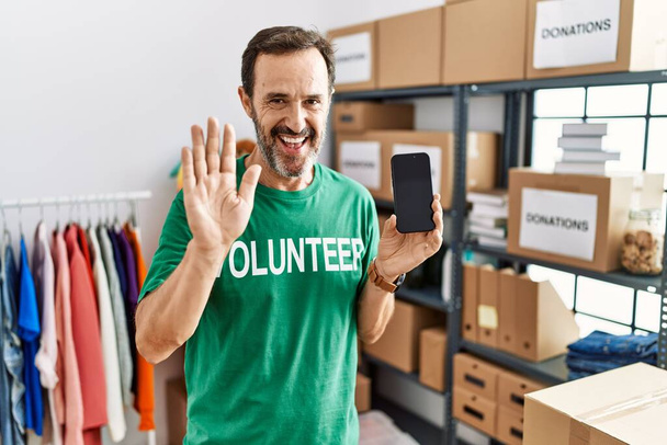 Middle age man with beard wearing volunteer t shirt holding smartphone waiving saying hello happy and smiling, friendly welcome gesture  - Fotoğraf, Görsel