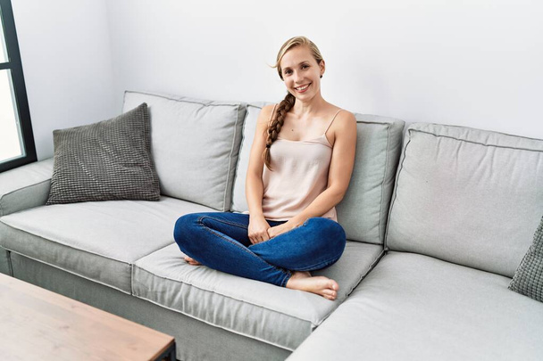 Νεαρή καυκάσια γυναίκα χαμογελά αυτοπεποίθηση κάθεται στον καναπέ στο σπίτι - Φωτογραφία, εικόνα