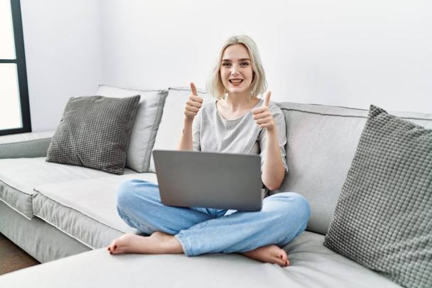 Молода біла жінка використовує ноутбук вдома, сидячи на дивані, схвалюючи позитивний жест рукою, великі пальці посміхаються і щасливі за успіх. жест переможця
.  - Фото, зображення