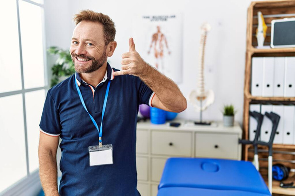 Middle age physiotherapist man working at pain recovery clinic smiling doing phone gesture with hand and fingers like talking on the telephone. communicating concepts.  - Foto, Imagen