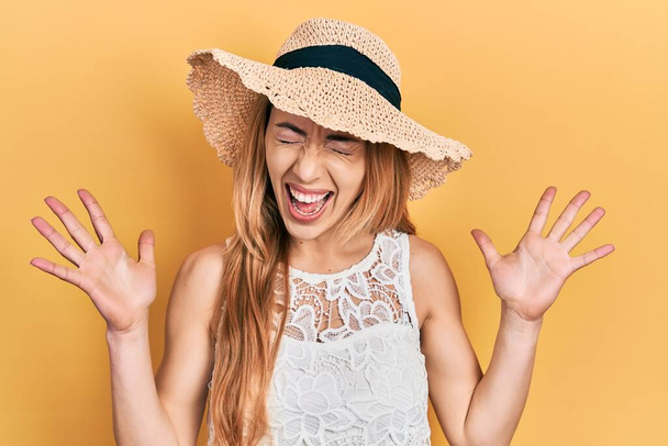 Young caucasian woman wearing summer hat celebrating mad and crazy for success with arms raised and closed eyes screaming excited. winner concept  - Foto, Imagen