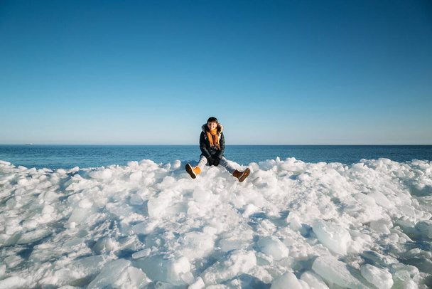 Young smiling woman sitting on a top of frozen sea ice blocks on a coast of the sea with a blue sky at the background, at frosty sunny day - Zdjęcie, obraz