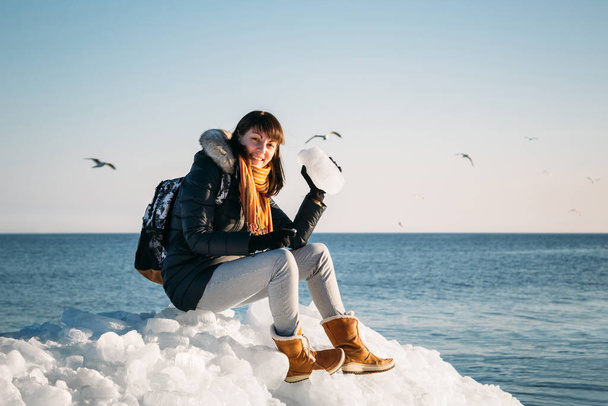 Молодая улыбающаяся женщина сидит на вершине морского льда блоков на побережье, держа сломанный кусок льда, с голубым морем и небом и на заднем плане - Фото, изображение