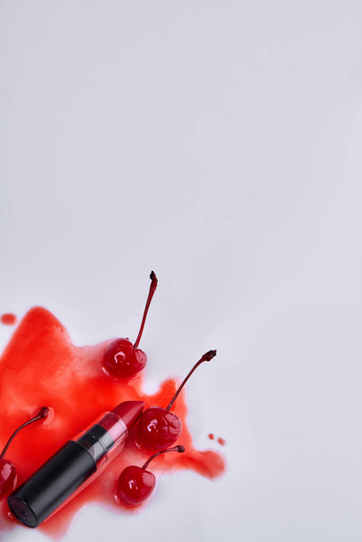 Vertical shot of lipstick and cherries on liquid stain. White background with copy space. - Foto, Imagem