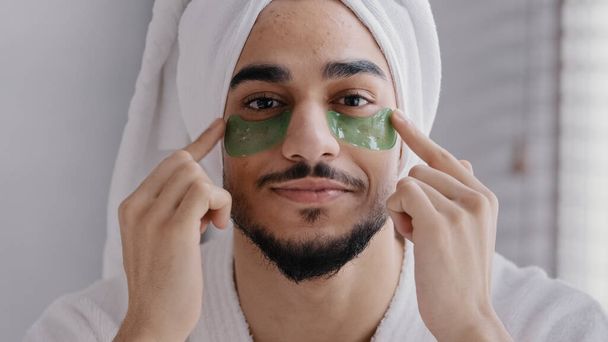 Front view portrait male face smiling indian arabian man wearing bath towel head glue sticky hydrogel eye-patches cosmetic procedures skin care guy looking at camera with collagen patches under eyes - Foto, afbeelding