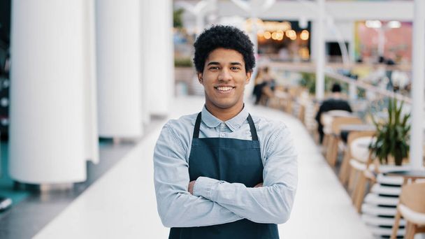 Confident African 30s restaurant worker biracial guy strong position standing indoors barman seller waiter in apron looking to camera successful cafe owner posing with crossed arms small business - Fotoğraf, Görsel