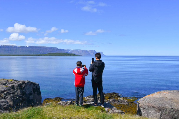 A boy and a young boy wear winter jacket with smartphone in hand while capturing a wide panorama of the sea and mountains in the background - Foto, afbeelding