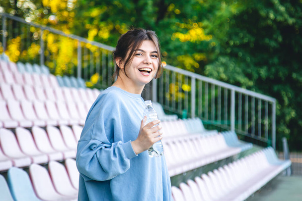 Attractive cheerful young woman sportswoman drinks water after training outdoors in the stadium. - Foto, Imagen