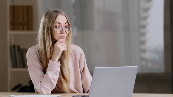 Caucasian millennial business woman female student girl writer journalist wearing glasses sitting at table at office home with laptop thinks idea imagination creative typing uses wireless computer app - Photo, Image