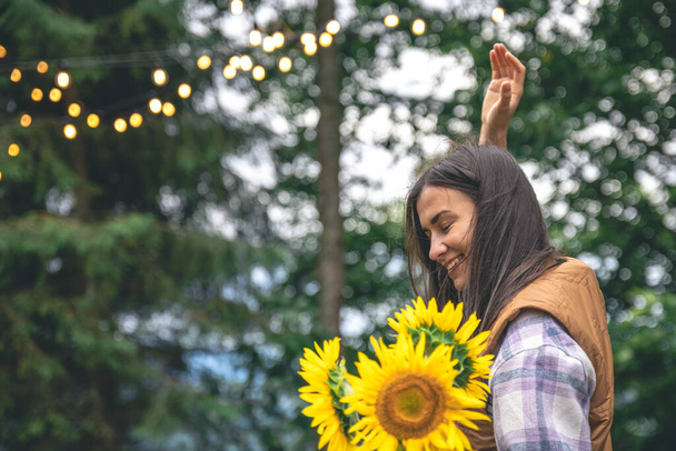 Nuori nainen, jolla on auringonkukkakimppu hämärtyneellä taustalla luonnossa bokeh-lampuilla. - Valokuva, kuva