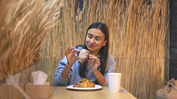Eine junge Frau im blauen Kapuzenpullover isst Croissants mit Kaffee in einem Café. - Foto, Bild