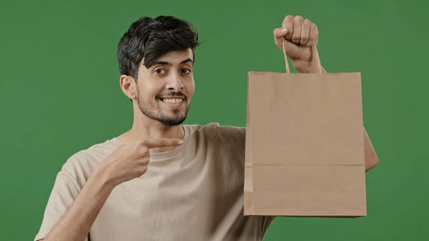 Portrait young smiling guy holding paper package pointing finger to shopping bag hispanic male buyer standing in studio on green background pleased happy man customer enjoying with successful purchase - Photo, Image